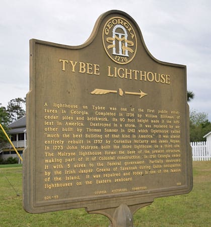 A historical marker for the Tybee Island Lighthouse, providing detailed information about its history, with the lighthouse and lush greenery visible in the background under natural lighting.