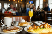 Cozy interior of Sunrise Restaurant on Tybee Island with wooden tables, plates of pancakes, omelets, shrimp and grits, coffee and mimosas, and sunlight streaming through windows in a lively breakfast scene.